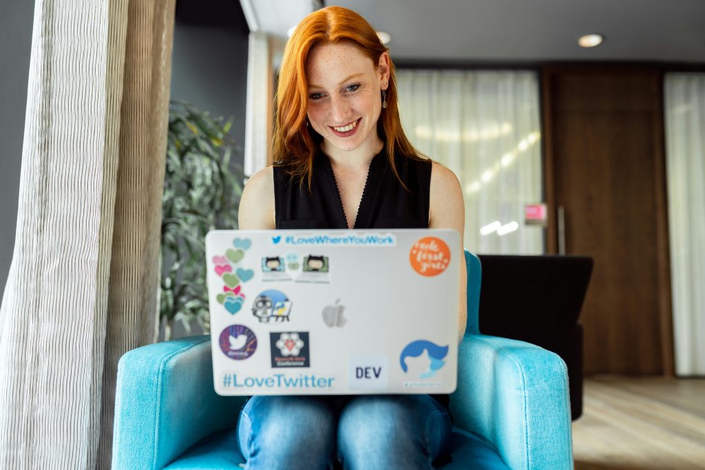 A cheerful young woman with red hair, working remotely on her laptop adorned with tech-related stickers. As remote work and digital skills gain popularity, coding and software development remain among the Top 9 High-Income Skills That Will Make You Money in 2025 by offering lucrative freelance and full-time job opportunities.