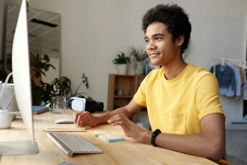 A young student wearing a bright yellow t-shirt sits at a modern wooden desk, attentively engaging with his computer screen. His expression is focused and enthusiastic, reflecting an immersive online learning experience. The workspace is well-lit, featuring a wireless keyboard, sticky notes, and a VR headset placed beside a glass of water. The background includes houseplants, a bookshelf, and neatly hung clothes, creating a comfortable home-study environment. This scene perfectly illustrates 10 Great Benefits Of Online Learning 2025, highlighting flexibility, accessibility, and the integration of advanced technology in virtual education.