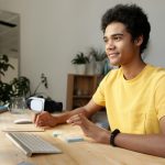 A young student wearing a bright yellow t-shirt sits at a modern wooden desk, attentively engaging with his computer screen. His expression is focused and enthusiastic, reflecting an immersive online learning experience. The workspace is well-lit, featuring a wireless keyboard, sticky notes, and a VR headset placed beside a glass of water. The background includes houseplants, a bookshelf, and neatly hung clothes, creating a comfortable home-study environment. This scene perfectly illustrates 10 Great Benefits Of Online Learning 2025, highlighting flexibility, accessibility, and the integration of advanced technology in virtual education.