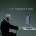 An elderly man with gray hair and a black sweater sits at a modern white desk, typing on a computer. The screen displays the words 'too old,' while a large projected text behind him reads 'no vacancies.' The dim lighting and minimalist setting emphasize the growing concern about how Artificial Intelligence (AI) is changing the job market, particularly in terms of hiring biases and automated decision-making. This thought-provoking image highlights AI’s role in employment discrimination and challenges older job seekers face in an evolving workforce.