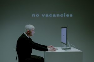 An elderly man with gray hair and a black sweater sits at a modern white desk, typing on a computer. The screen displays the words 'too old,' while a large projected text behind him reads 'no vacancies.' The dim lighting and minimalist setting emphasize the growing concern about how Artificial Intelligence (AI) is changing the job market, particularly in terms of hiring biases and automated decision-making. This thought-provoking image highlights AI’s role in employment discrimination and challenges older job seekers face in an evolving workforce.